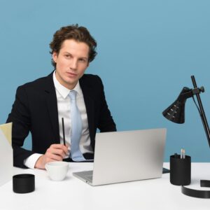 A focused businessman in a suit works at his tidy desk with a laptop, plant, and lamp.